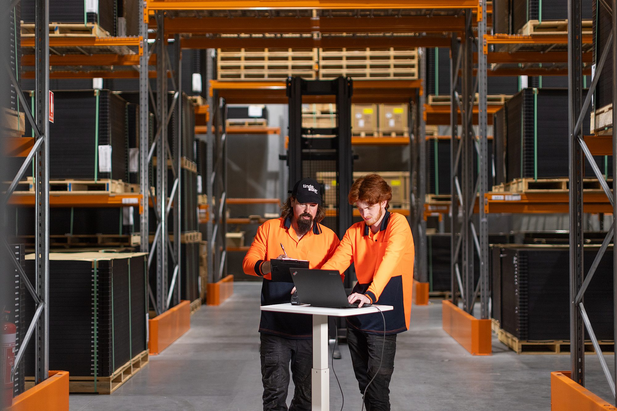 Two tondo solar workers wearing orange high vis are in a warehouse. One is taking notes and another is looking at a laptop.